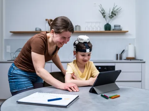 child sitting at desk in front of a tablet wearing a brainhero device with an adult behind